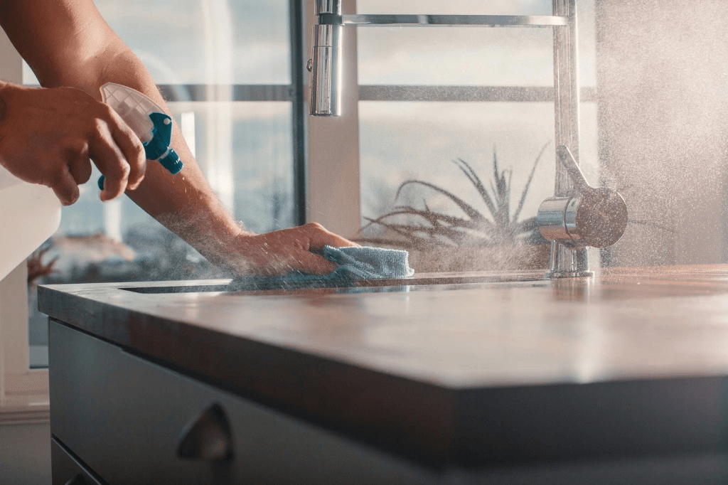 Hands spraying and wiping a kitchen countertop in a sunlit kitchen
